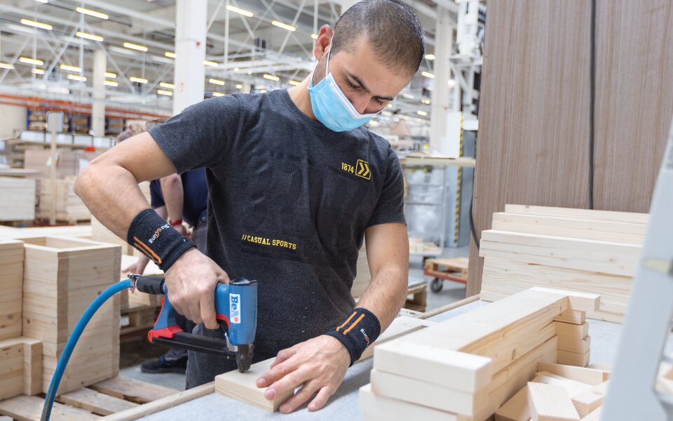 Mitarbeiter in Produktion mit Mundschutz und Bohrer an Holzteilen