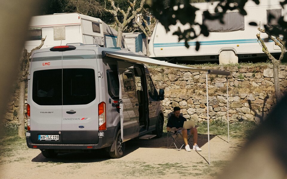 LMC Camper vor einer Mauer, Mann sitzt davor am Laptop arbeitend im Campingstuhl