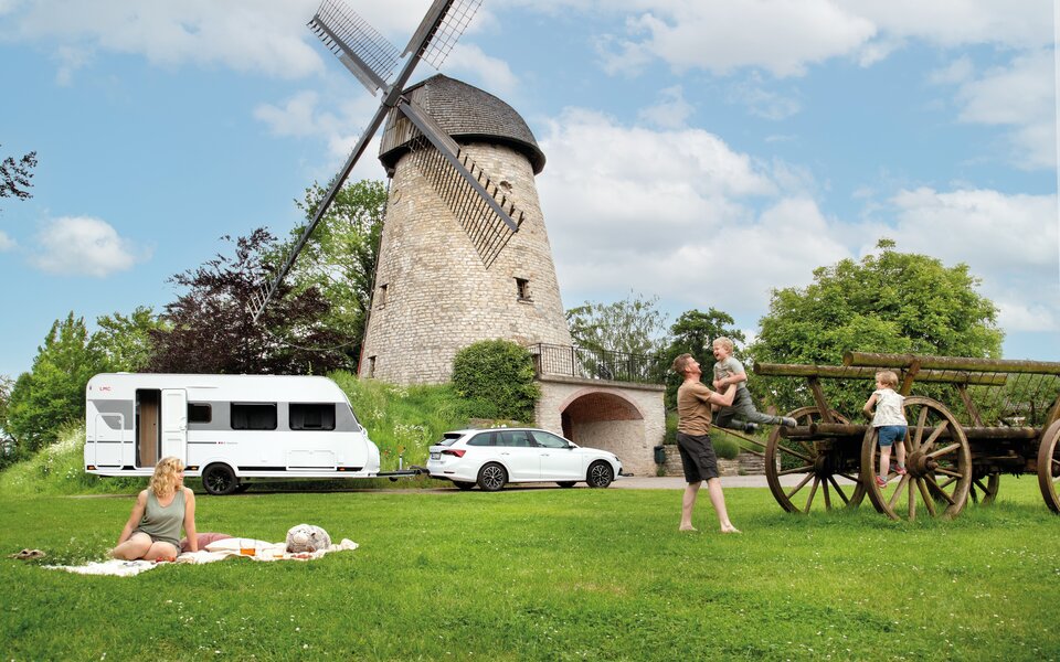 Familie mit zwei Kindern auf großer Wiese, Frau sitzt auf Decke, Mann und Kinder spielen, im Hintergrund Windmühle und LMC Wohnwagen