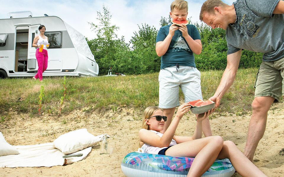 Familie am Strand mit Badeutensilien und Schwimmreifen, Wassermelone essend, im Hintergrund ein LMC Wohnwagen