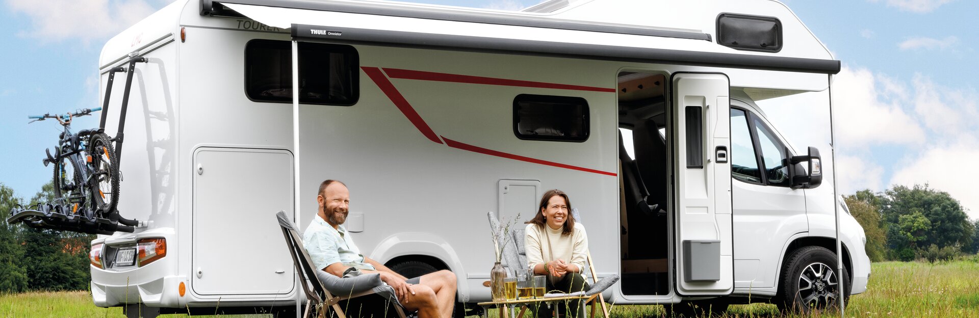 Ehepaar auf Campingstühlen sitzend vor LMC Wohnwagen auf grüner Wiese